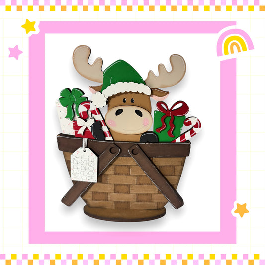 A wooden moose wearing a Santa hat is sitting in a brown wicker basket filled with Christmas gifts, including presents and candy canes.