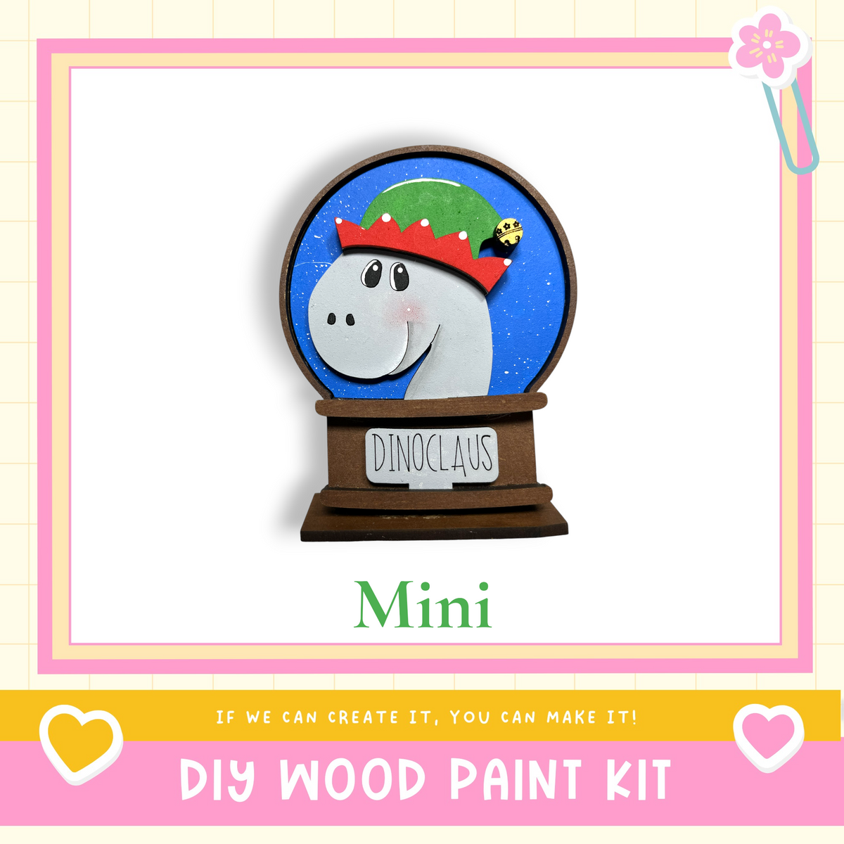 a wooden snow globe with a dinosaur wearing a red hat and the word "Dinoclawus" written on it. The snow globe is surrounded by a pink border and a heart symbol. The image also includes text that says "Mini DIY WOOD PAINT KIT" and "If we can create it, you can make it!"