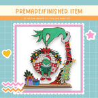 The image is a collage featuring a hand holding a Christmas ornament with a green frog wearing a Santa hat and reading a book. The ornament is surrounded by a wreath, presents, and a Christmas tree. The background is a light blue color with a grid pattern.