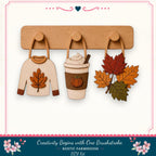 a wooden hanger with three decorative items hanging on it: a sweater with a leaf design, a coffee cup with a pumpkin and whipped cream design, and a leaf arrangement.