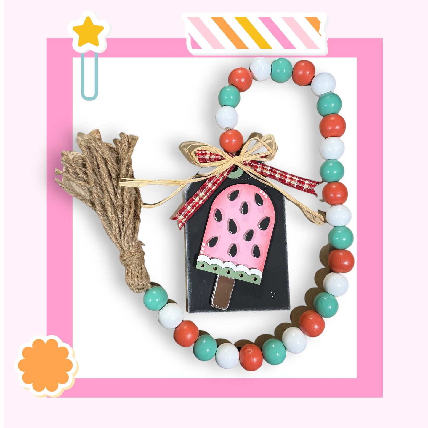 a pink and white frame with a beaded necklace, a tassel, and a small wooden block with a pink and black design.