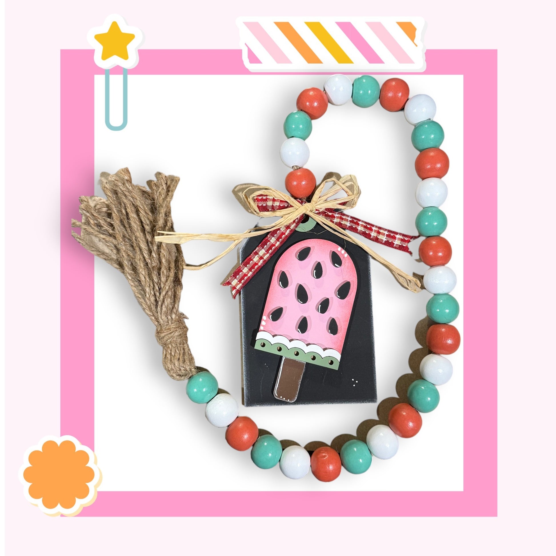a pink and white frame with a beaded necklace, a tassel, and a small wooden block with a pink and black design.