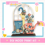 The image is a colorful and whimsical DIY wood paint kit featuring a wooden window with a flower arrangement, a pair of blue rain boots, and a pink watering can.