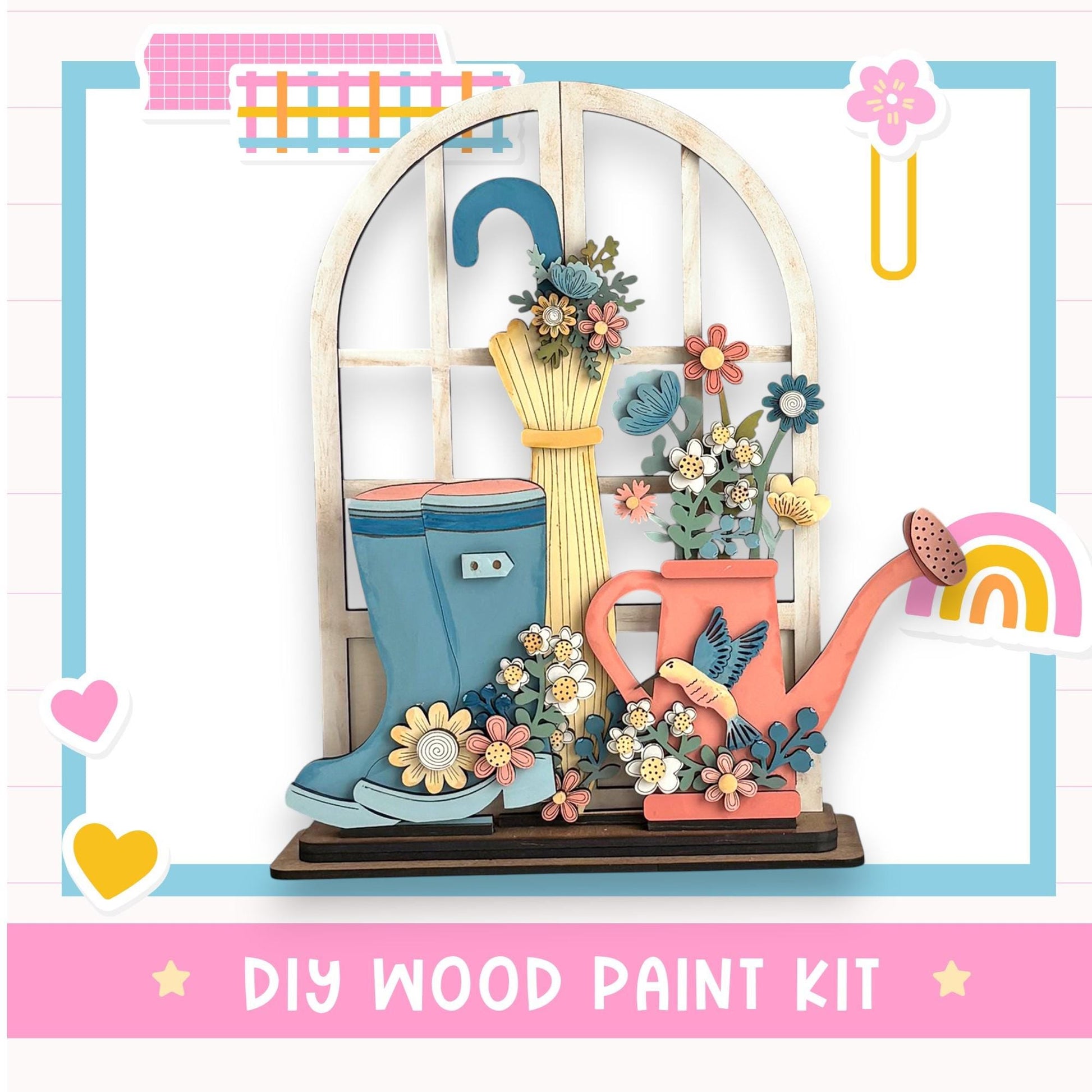 The image is a colorful and whimsical DIY wood paint kit featuring a wooden window with a flower arrangement, a pair of blue rain boots, and a pink watering can.