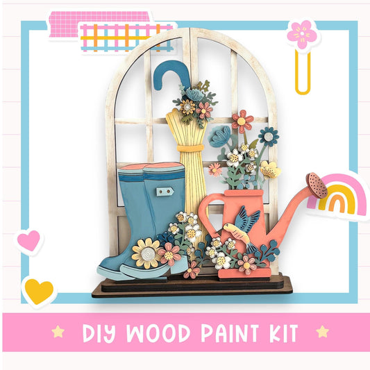 The image is a colorful and whimsical DIY wood paint kit featuring a wooden window with a flower arrangement, a pair of blue rain boots, and a pink watering can.