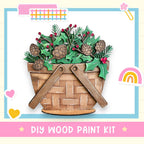 A DIY wood paint kit featuring a wooden basket filled with pine cones, holly leaves, and berries, with a rainbow and hearts in the background.