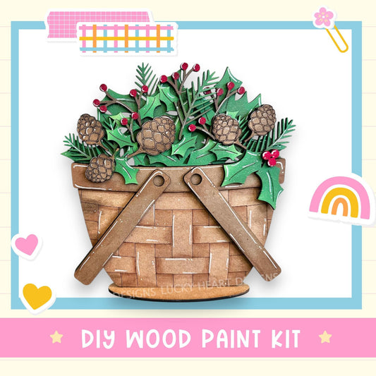 A DIY wood paint kit featuring a wooden basket filled with pine cones, holly leaves, and berries, with a rainbow and hearts in the background.