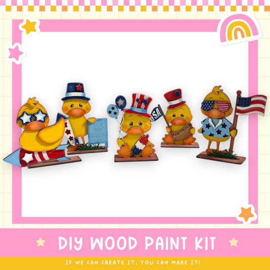 a DIY wood paint kit with five cheerful duck figurines wearing patriotic hats and holding American flags. The kit appears to be made of wood and is designed for painting.