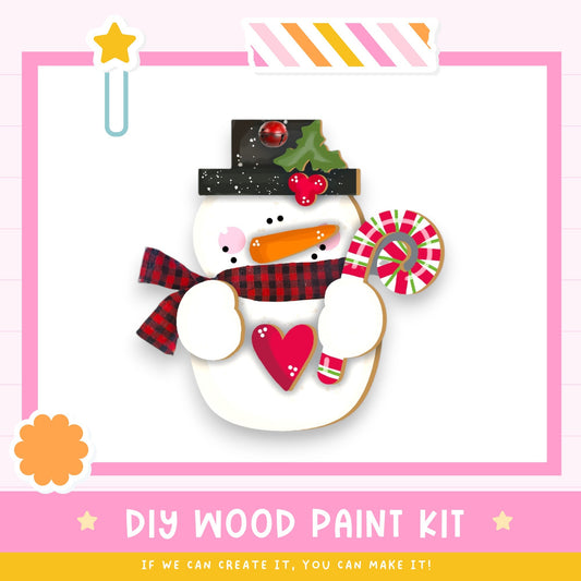 A snowman made from wood, wearing a hat, scarf, and holding a candy cane, is the central focus of the image. The background is a pink and white color scheme, and there is text at the bottom of the image that reads "DIY WOOD PAINT KIT" and "IF WE CAN CREATE IT, YOU CAN MAKE IT!"