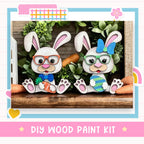 two white rabbits wearing glasses and holding carrots, sitting on a wooden surface. The rabbits are surrounded by a wreath of greenery and a rainbow-colored flower. The image also includes text that reads "DIY WOOD PAINT KIT" and "Easter Bunny".
