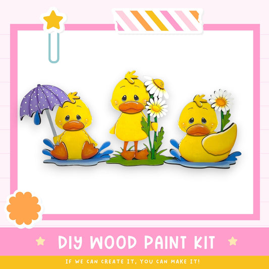 three yellow duck figures sitting on a grassy surface, each holding an umbrella and holding flowers. The image is a collage of multiple images, and the text below the image reads "DIY WOOD PAINT KIT - IF WE CAN CREATE IT, YOU CAN MAKE IT!"