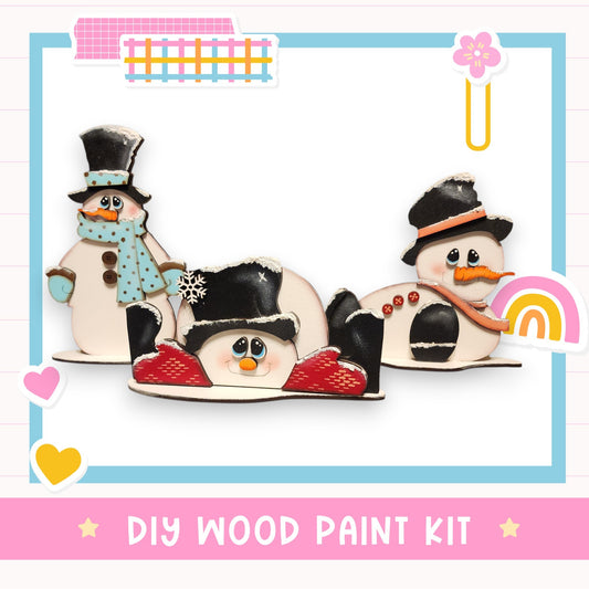 a DIY wood paint kit with three wooden snowmen, each wearing a hat and scarf, and holding a snowflake.
