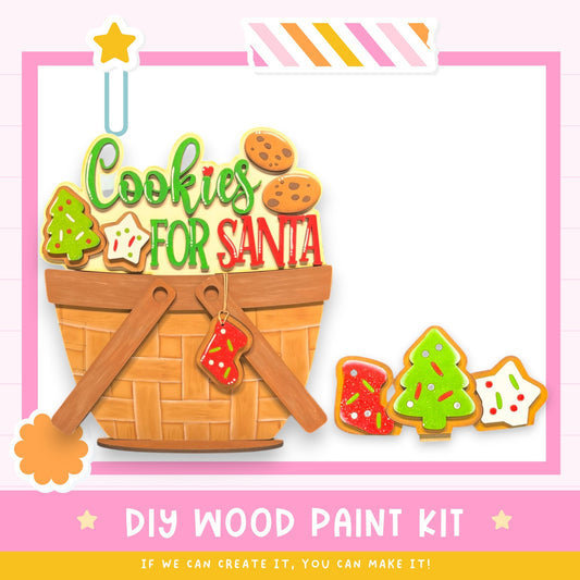 The image is a colorful and festive DIY wood paint kit for creating a "Cookies for Santa" themed decoration. The kit includes a wooden basket filled with various cookie shapes, as well as a wooden paintbrush and a wooden stick. The kit is designed to be used by children to create their own "Cookies for Santa" decorations.