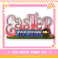 The image is a colorful Easter-themed sign with the text "Easter Egg Hunt" and a basket of Easter eggs. The sign is surrounded by a pink and yellow border with stars and a rainbow. The image also includes the text "DIY WOOD PAINT KIT" and "IF WE CAN CREATE IT, YOU CAN MAKE IT!"