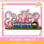 The image is a colorful Easter-themed sign with the text "Easter Egg Hunt" and a basket of Easter eggs. The sign is surrounded by a pink and yellow border with stars and a rainbow. The image also includes the text "DIY WOOD PAINT KIT" and "IF WE CAN CREATE IT, YOU CAN MAKE IT!"