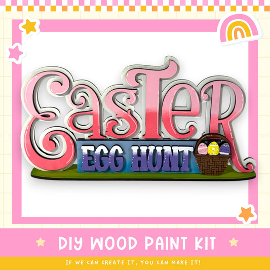 The image is a colorful Easter-themed sign with the text "Easter Egg Hunt" and a basket of Easter eggs. The sign is surrounded by a pink and yellow border with stars and a rainbow. The image also includes the text "DIY WOOD PAINT KIT" and "IF WE CAN CREATE IT, YOU CAN MAKE IT!"