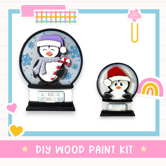 two snow globes, each containing a penguin character, set against a white background. The text "DIY WOOD PAINT KIT" is displayed at the bottom of the image.