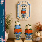 a creative DIY kit for decorating with pumpkins, including a welcome sign and decorative planters. The kit includes pumpkins in various colors, such as orange, white, and blue, as well as decorative elements like ribbons and flowers. The pumpkins are stacked in different sizes and placed in vases, creating a visually appealing display. The kit is set against a backdrop of a wall with a framed picture of pumpkins, adding to the festive atmosphere.