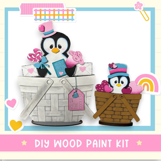 The image is a collage featuring two wooden toy penguins, one larger and one smaller, sitting in baskets and holding various items such as hearts, a book, and a rainbow. The image also includes a sign that reads "DIY WOOD PAINT KIT" and a pink background with a heart and a rainbow.