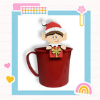 A red mug with a cartoon elf figure holding a gift box on top, set against a colorful background with various decorative elements.