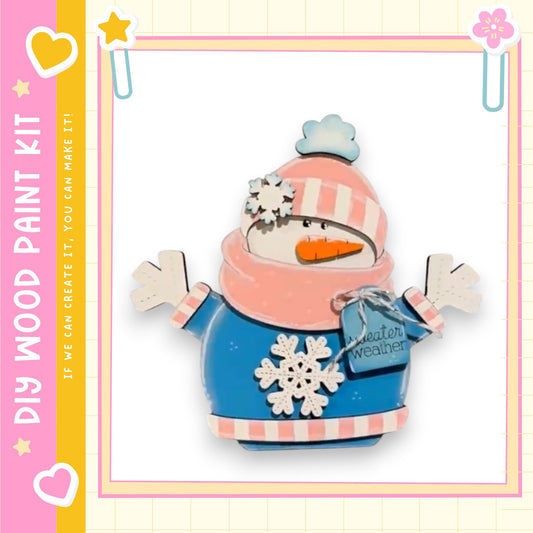 A wooden snowman kit with a blue and pink outfit, a carrot nose, and a snowflake headband, set against a pink and yellow background with a heart and star motif.