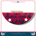 a red sign with the text "Sweet land of Liberty" written on it, surrounded by a decorative border with pink and blue floral elements.