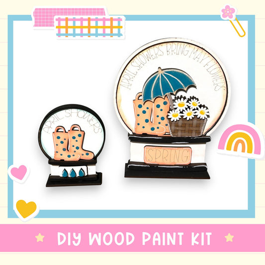 The image is a DIY wood paint kit featuring a wooden snow globe with a blue umbrella, flowers, and a rainbow, along with a smaller snow globe with a pair of boots.