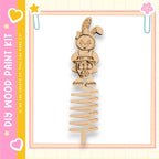 A wooden Easter bunny toy is shown on a spring-like structure, surrounded by a pink and yellow border.