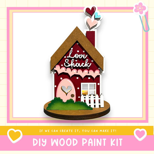 a wooden house-shaped object with a heart-shaped window and a heart-shaped door, surrounded by hearts and hearts. The text below the image reads "DIY WOOD PAINT KIT" and "If we can create it, you can make it!"