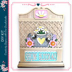 The image is a wooden sign with a decorative design, featuring a teapot, flowers, and a heart. The sign has the text "Spring Blossoms" written on it.