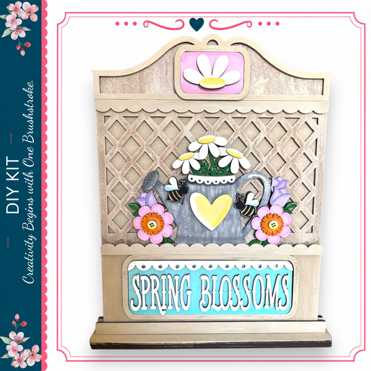 The image is a wooden sign with a decorative design, featuring a teapot, flowers, and a heart. The sign has the text "Spring Blossoms" written on it.