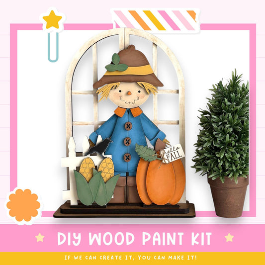 a DIY wood paint kit for creating a scarecrow, with a wooden scarecrow figure holding a pumpkin and corn cobs, and a potted plant nearby.