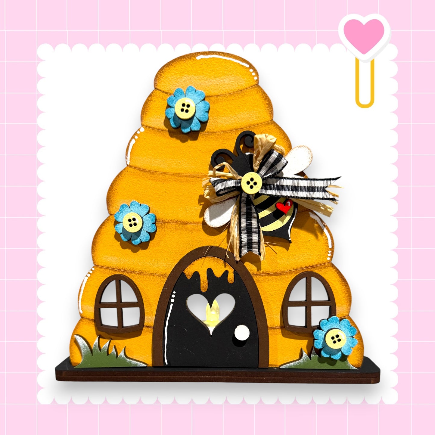 A whimsical, handcrafted wooden bee hive with a black door and windows, adorned with blue and yellow flowers, and a heart-shaped decoration.