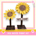 two wooden sunflower sculptures, each with a wooden sign attached to them. The sign says "WELCOME". The sculptures are placed on a wooden stand, and there is a DIY wood paint kit visible in the image.
