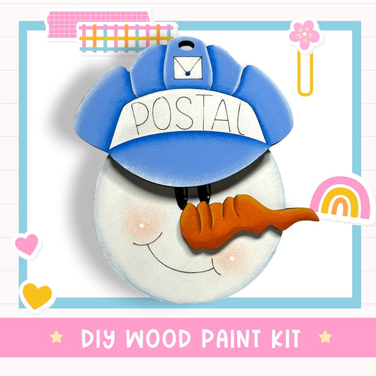 A wooden postcard-style snowman with a blue hat and a carrot nose, surrounded by various colorful stickers and a pink banner with the text "DIY WOOD PAINT KIT".
