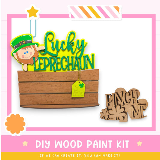 The image is a colorful and playful DIY wood paint kit featuring a leprechaun, a wooden sign, and a wooden sign with the phrase "Pinch Me". The kit appears to be designed for children to create their own wooden signs and decorations.
