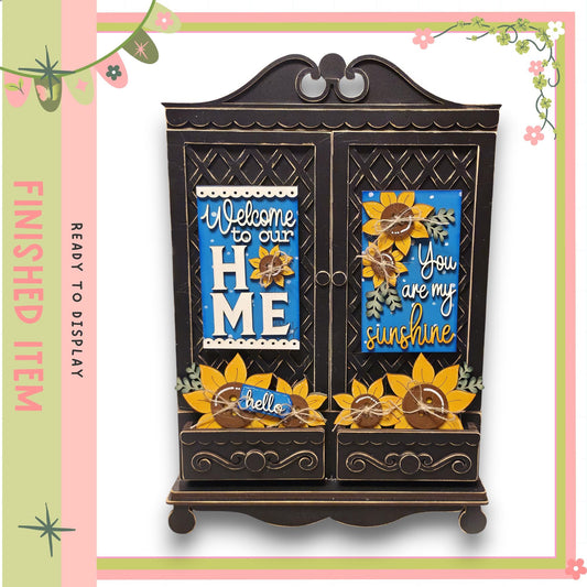 A decorative wooden cabinet with a sign that reads "Welcome to our home" and another sign that says "You are my sunshine". The cabinet has a floral design and is surrounded by a pink and green border.