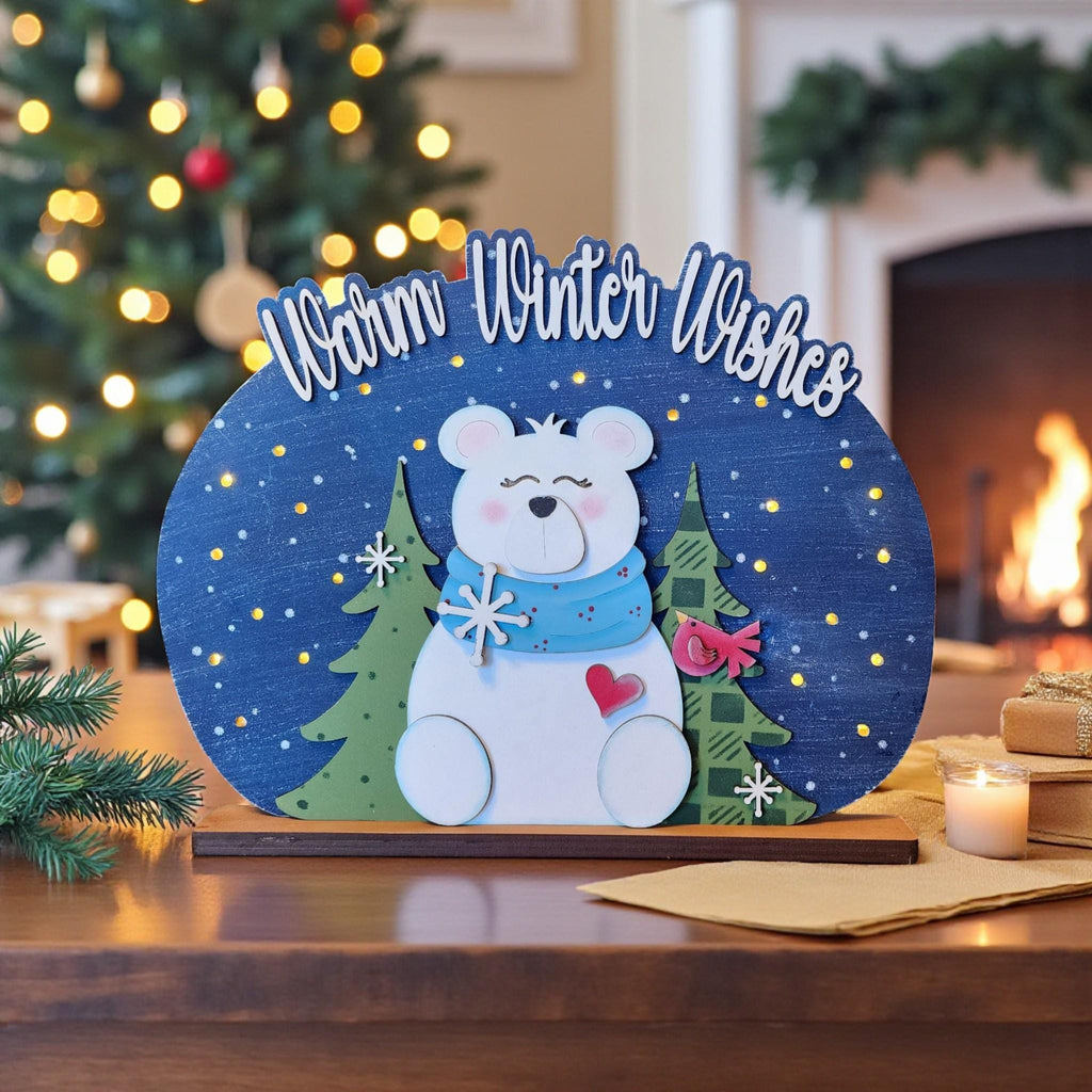 A wooden sign with a polar bear and snowflakes, surrounded by a winter scene, sits on a table with a lit candle and other holiday decorations in the background.