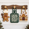 three wooden ornaments hanging on a wooden board, each depicting a different holiday-themed character or house.