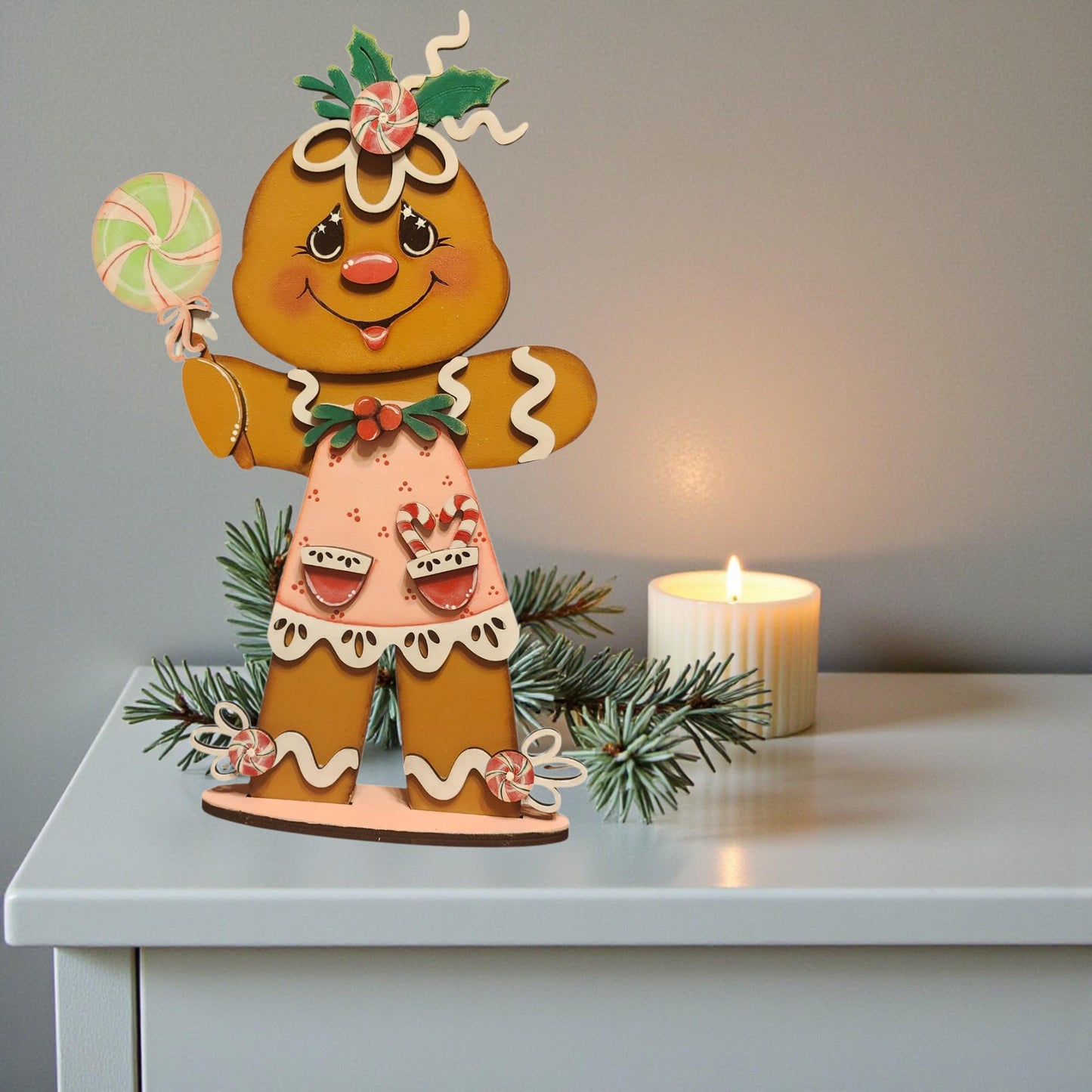 A gingerbread man figurine holding a lollipop and surrounded by pine branches and a lit candle on a white surface.