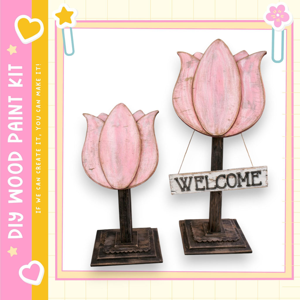 two wooden flower-shaped sculptures, each with a wooden sign attached to them. The sign reads "WELCOME". The sculptures are placed on a wooden base, and the background is a light pink color with a yellow border.