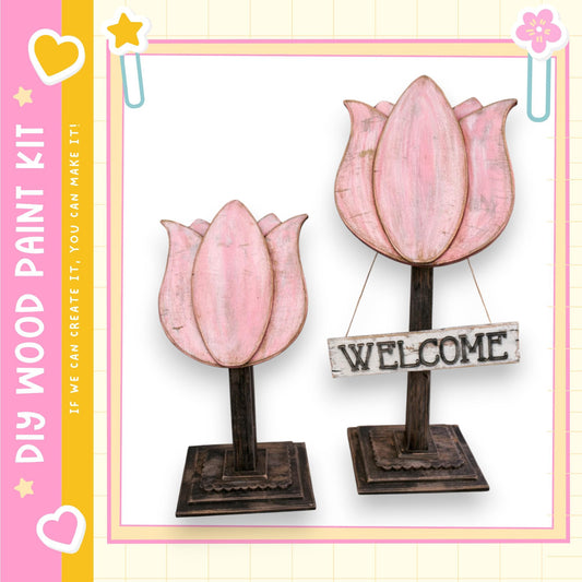 two wooden flower-shaped sculptures, each with a wooden sign attached to them. The sign reads "WELCOME". The sculptures are placed on a wooden base, and the background is a light pink color with a yellow border.