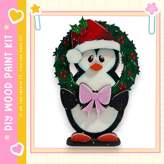 A wooden penguin wearing a Santa hat and holding a bow, standing in front of a wreath.