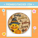 a wooden sign with a duck wearing a witch's hat and holding a pumpkin, surrounded by autumn leaves.