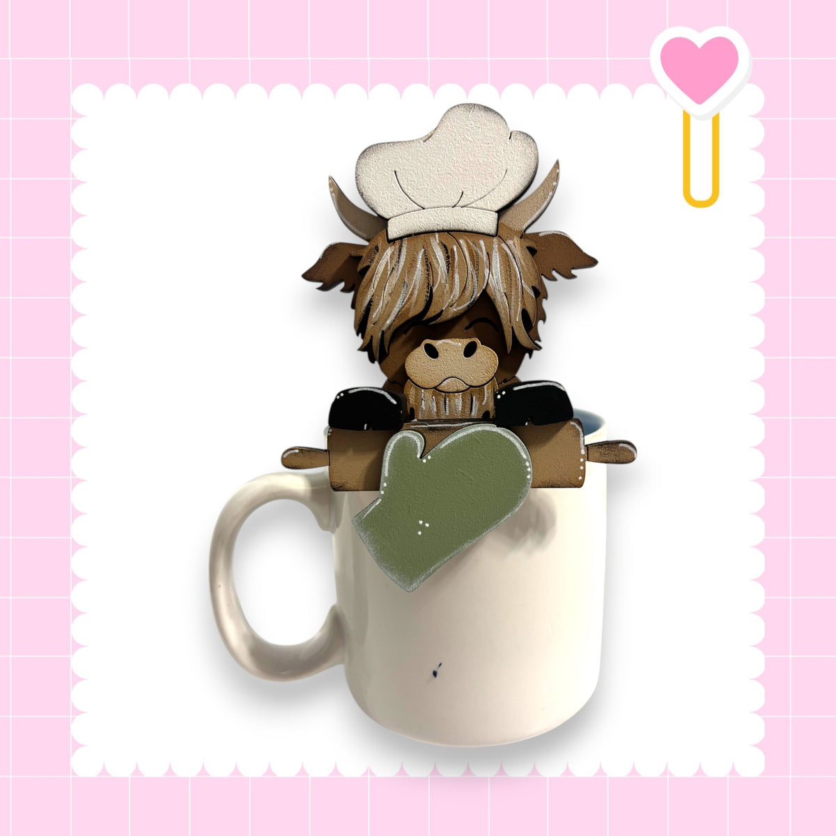 A cartoon cow wearing a chef's hat and holding a rolling pin is sitting inside a white mug, set against a pink background with a heart shape.