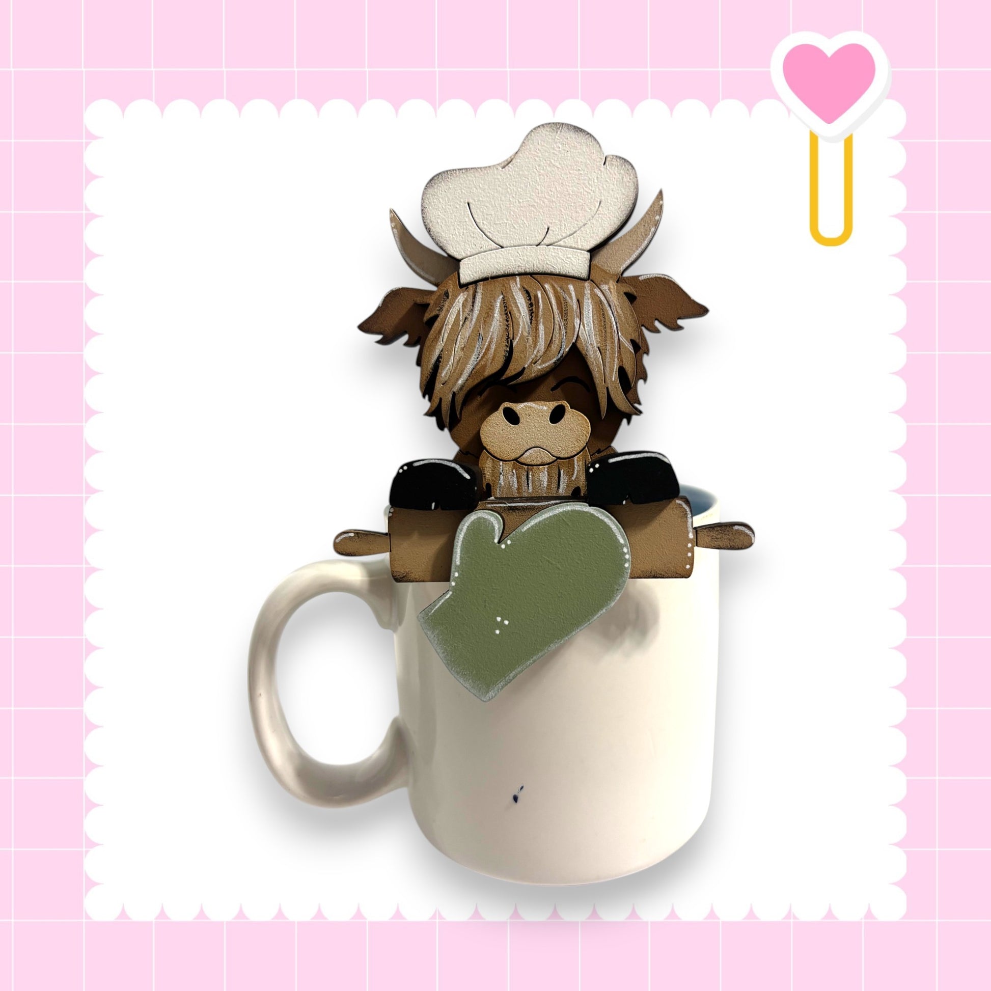 A cartoon cow wearing a chef's hat and holding a rolling pin is sitting inside a white mug, set against a pink background with a heart shape.