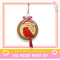 a DIY wood paint kit with a wooden ornament hanging from a string, depicting a red cardinal bird. The kit includes a heart and a snowflake ornament, and the text encourages creating the kit if one can.