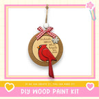 a DIY wood paint kit with a wooden ornament hanging from a string, depicting a red cardinal bird. The kit includes a heart and a snowflake ornament, and the text encourages creating the kit if one can.