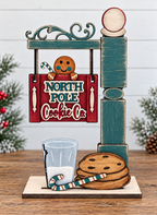 A wooden sign for "North Pole Cookie Co.", featuring a gingerbread man and a cookie, is displayed on a wooden stand with a glass of milk and a cookie.
