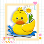 A yellow duck sitting in a puddle, with a white flower and a pink heart decoration nearby.
